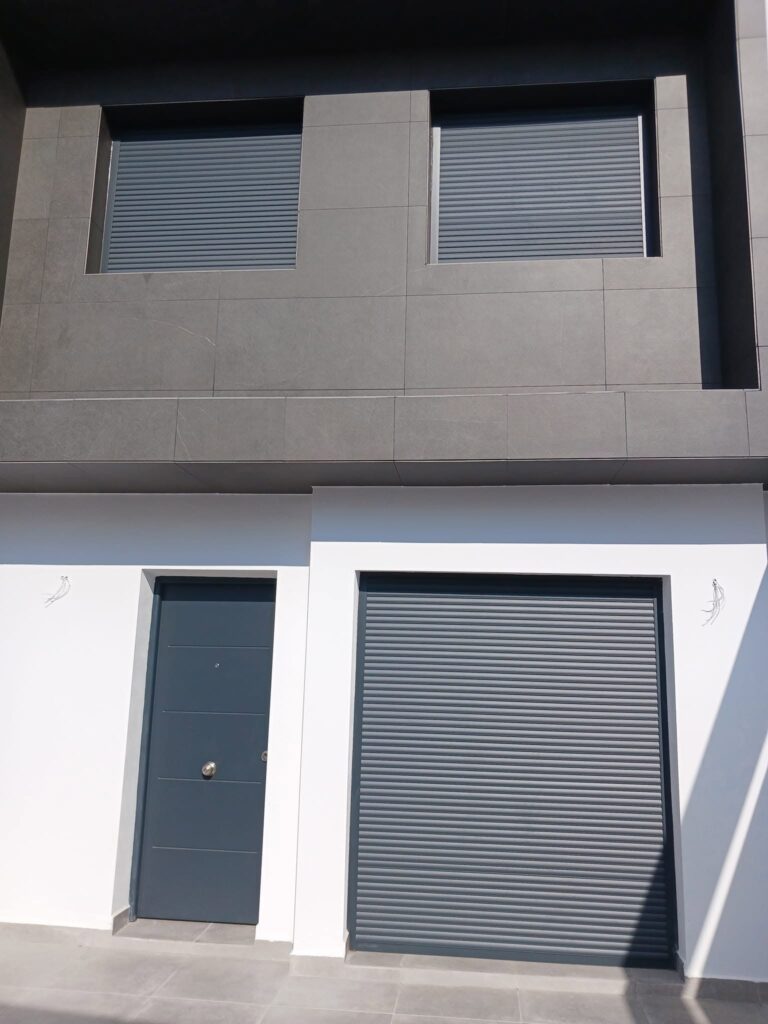 puerta-y-persianas-metalicas-vivienda Fachada de vivienda con puerta de entrada y persianas enrollables metálicas en color gris oscuro